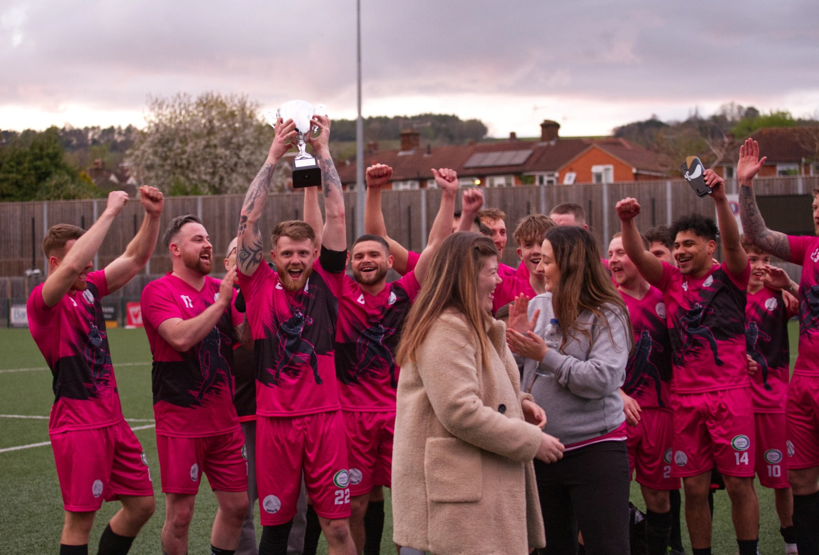 Football team in pink and black kits celebrating with a trophy